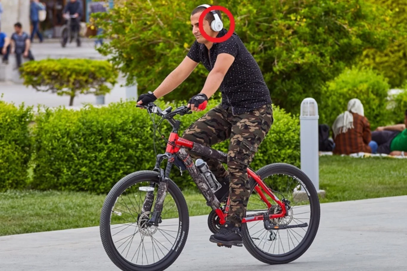 Zdecydowanie najlepszym sposobem będzie zmiana utworów lub głośności gestami, ponieważ korzystanie z telefonu lub innego urządzenia elektrycznego podczas jazdy jest zabronione. Mężczyzna w czarnej koszulce i spodniach moro jedzie czerwonym rowerem, ma na uszach białe słuchawki, w tle zielone krzewy i trawnik, na którym siedzą ludzie.