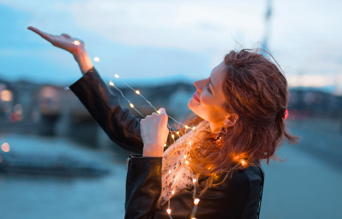 Andrzejki to idealny moment na wróżby. Sprawdź, co cię czeka w miłości Kobieta w ciemnej kurtce, otulona świecącymi lampkami LED, uśmiecha się i spogląda w górę, stojąc na tle rozmytego pejzażu miejskiego o zmierzchu.