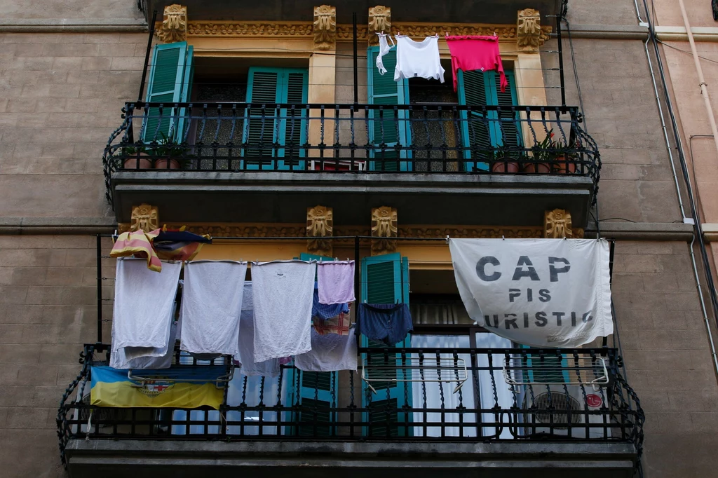 Barcelona. Baner wywieszony podczas protestów przeciwko wynajmowaniu mieszkań turystom Balkon z rozwieszonym praniem, zielone okiennice, baner z napisem „CAP PIS TURISTIC” oraz suszące się ubrania i ręczniki. Elewacja kamienicy w tle.
