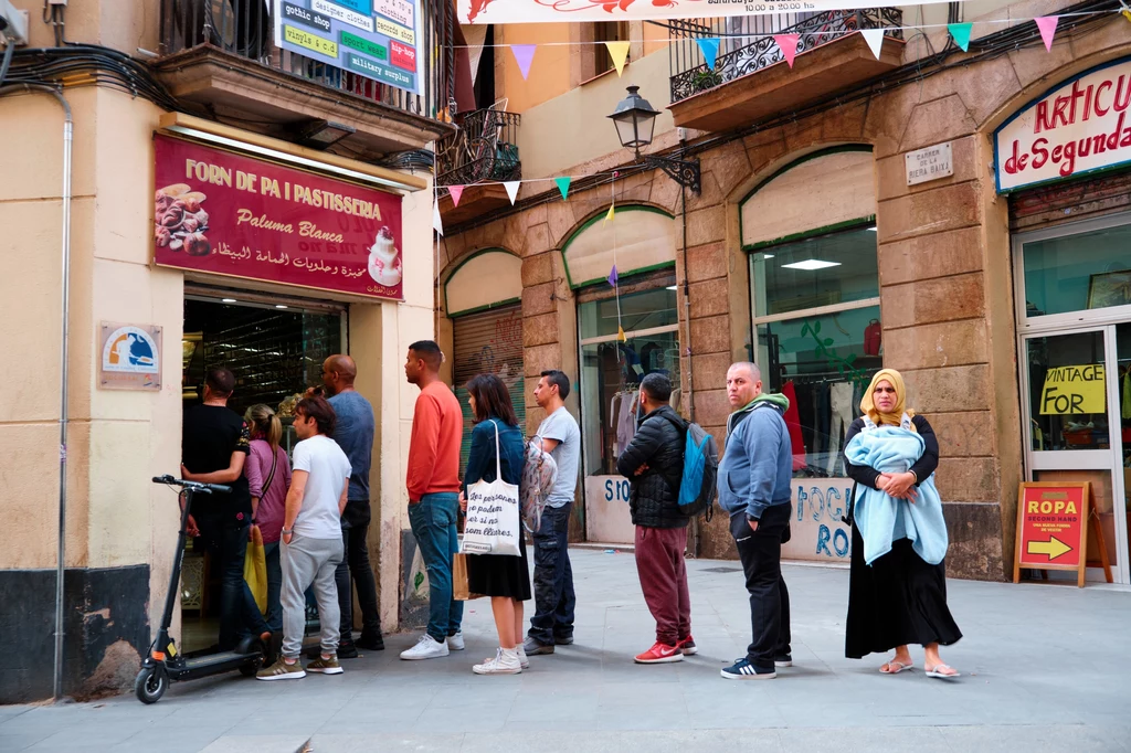 Barcelona Ludzie ustawieni w kolejce przed piekarnią na ulicy miasta, niektórzy rozmawiają, inni czekają spokojnie, kobieta z dzieckiem na rękach stoi na końcu grupy, otoczenie miejskie z witrynami i banerami.