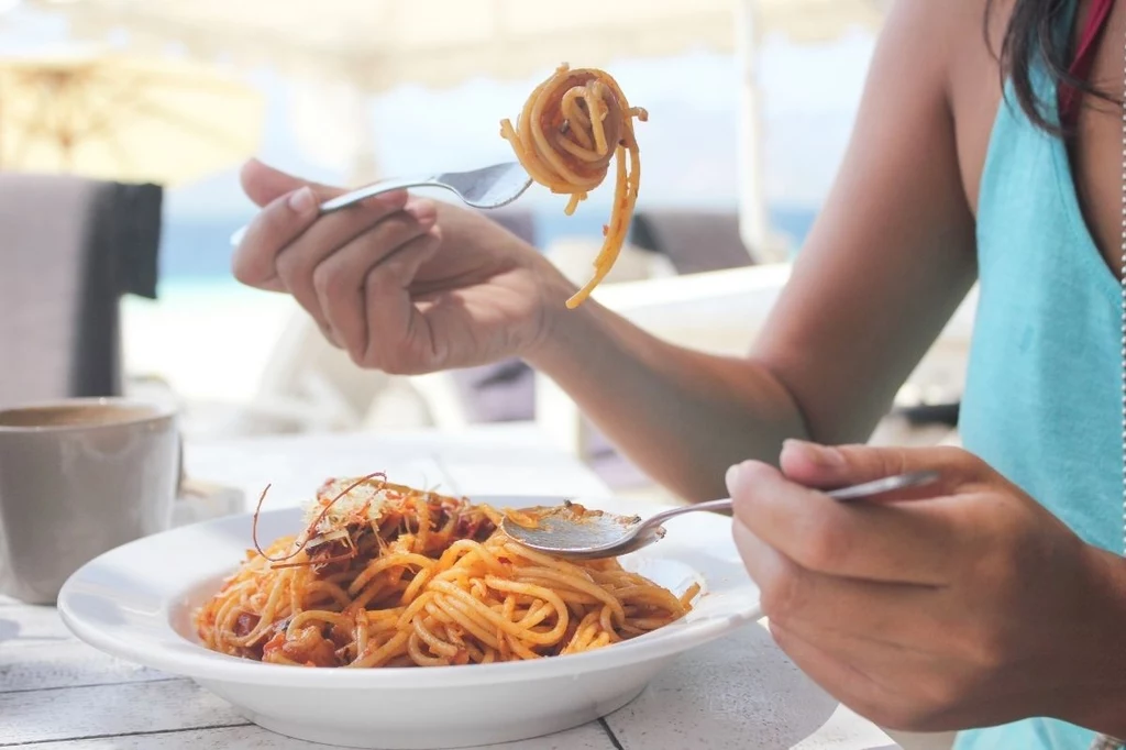 Naukowcy przekonują, że jedzenie makaronu zapewnia nam poczucie szczęścia Talerz spaghetti z sosem pomidorowym, osoba trzyma widelce i nakręca makaron, obok kubek z napojem, jasne, wakacyjne otoczenie.