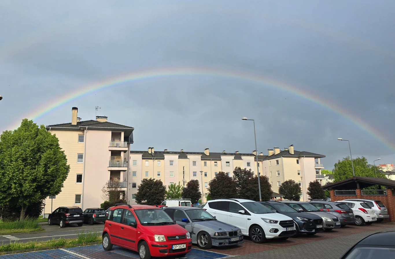 Jak powstaje tęcza i ile ma kolorów? Kolorowa tęcza rozpościera się nad wielopiętrowymi blokami mieszkalnymi, na pierwszym planie parking z szeregiem zaparkowanych samochodów, z boku widoczne drzewa i latarnie uliczne, niebo częściowo zachmurzone.