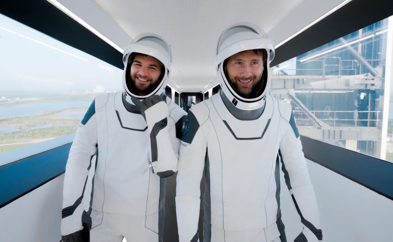 Polak leci w kosmos. Kiedy start i co warto o nim wiedzieć? Dwóch uśmiechniętych astronautów w nowoczesnych białych skafandrach kosmicznych spaceruje przeszklonym korytarzem przy platformie startowej, w tle nowoczesna infrastruktura i fragment panoramy krajobrazu.
