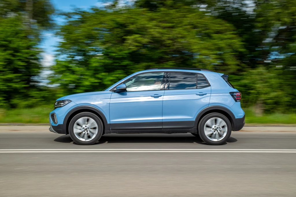 Volkswagen T-Cross to jeden z najpopularniejszych modeli niemieckiego producenta Jasnoniebieski samochód typu SUV jedzie po drodze asfaltowej na tle zielonych drzew, ruch pojazdu uchwycono w lekkim rozmyciu wskazującym na prędkość.