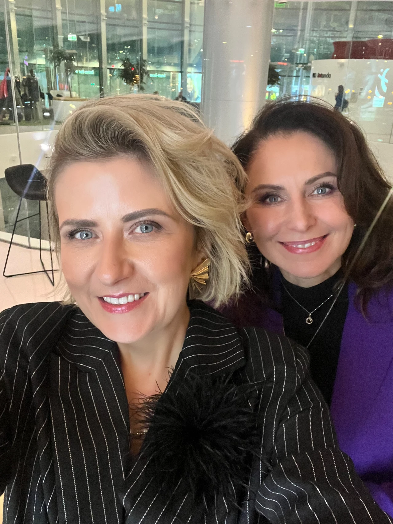 Anna Janiczek i Katarzyna Plewa, założycielki Fundacji The Best Age Dwie kobiety pozujące do selfie w nowoczesnym wnętrzu z przeszklonymi ścianami oraz elementami wystroju takimi jak wysoki słupek i widoczne kawiarniane stoliki w tle, obie uśmiechnięte, ubrane elegancko w marynarki.