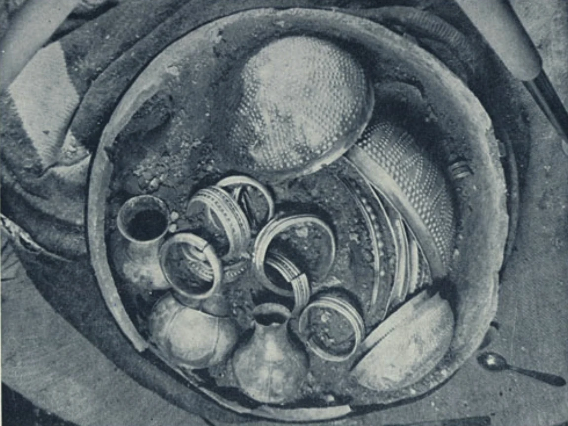 Zbiór dawnych naczyń, bransolet i mis wykonanych z metalu, pokazanych w okrągłym naczyniu, wokół których widoczne są delikatne warstwy ziemi.