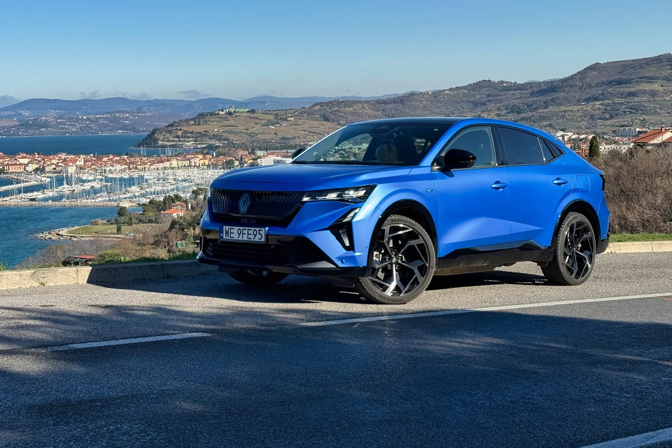Renault Rafale w wersji plug-in dysponuje napędem na 4 koła i mocą 300 KM Nowoczesny, niebieski samochód osobowy zaparkowany przy asfaltowej drodze z widokiem na malowniczą marinę, miasteczko i wzgórza w tle, w pogodny dzień.
