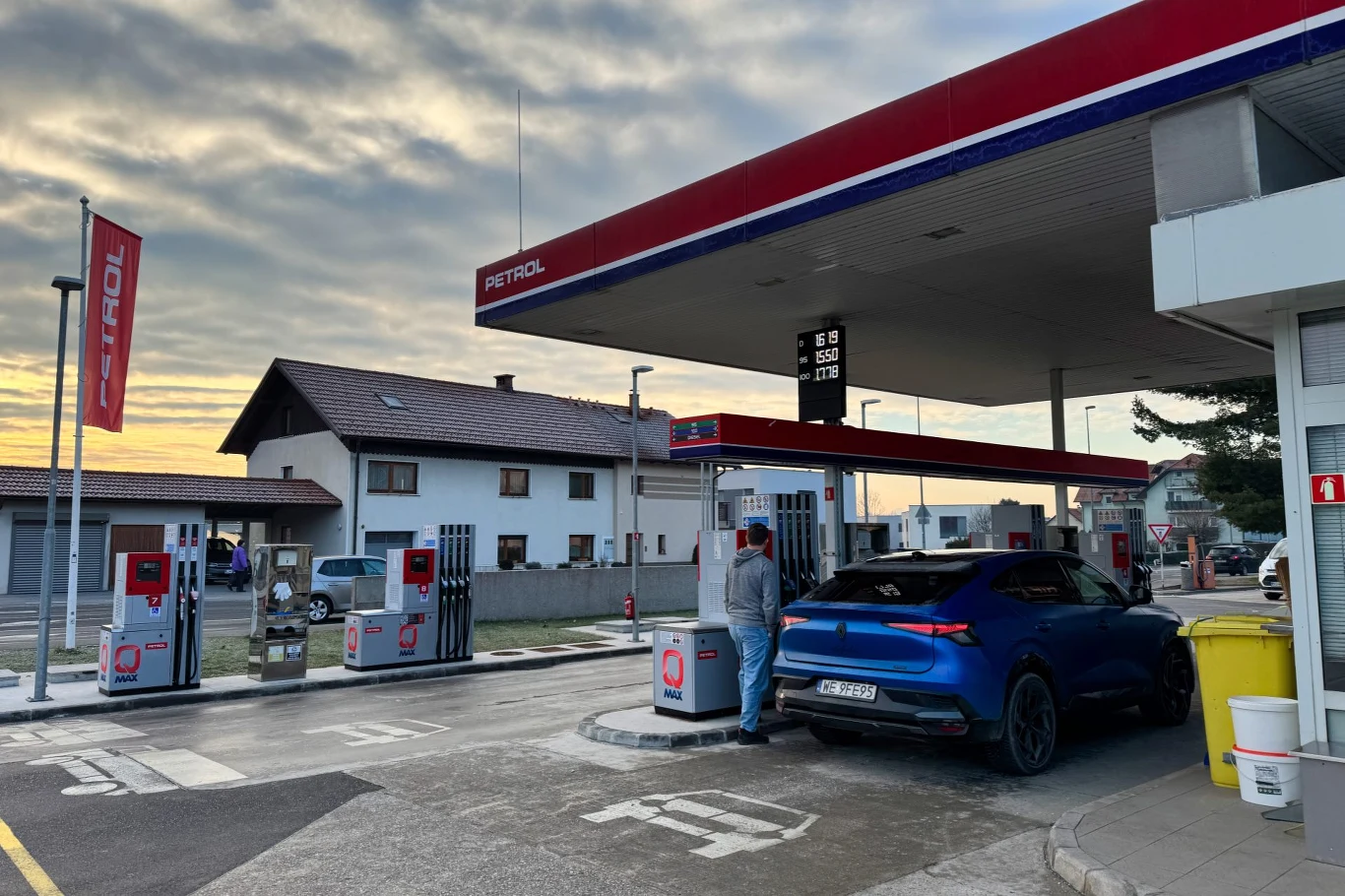 Dzięki hybrydowemu napędowi i dużej baterii Rafale PHEV nie było częstym gościem stacji paliw Stacja paliw z nowoczesnym dystrybutorem, na pierwszym planie niebieski samochód ładowany przez stojącego przy nim mężczyznę, w tle budynek mieszkalny oraz zachmurzone niebo.