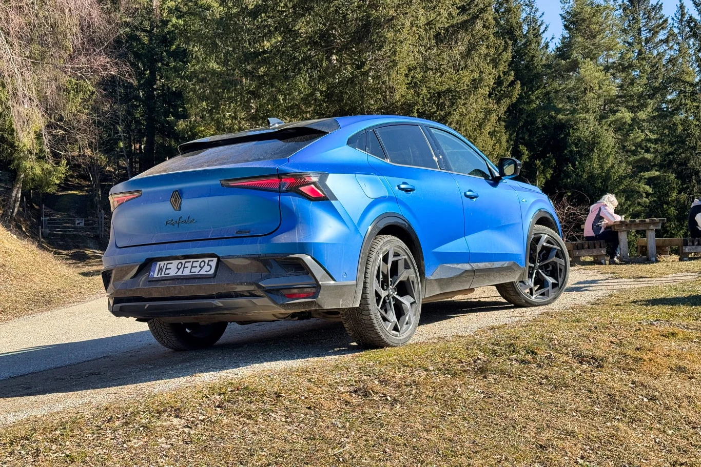 Satynowy lakier i dużo elementów z fortepianowej czerni sprawiały, że każda drobina kurzu była widoczna z daleka Niebieski SUV marki MG4 Electric zaparkowany na poboczu drogi leśnej, w tle drzewa iglaste i osoby siedzące przy stole piknikowym.