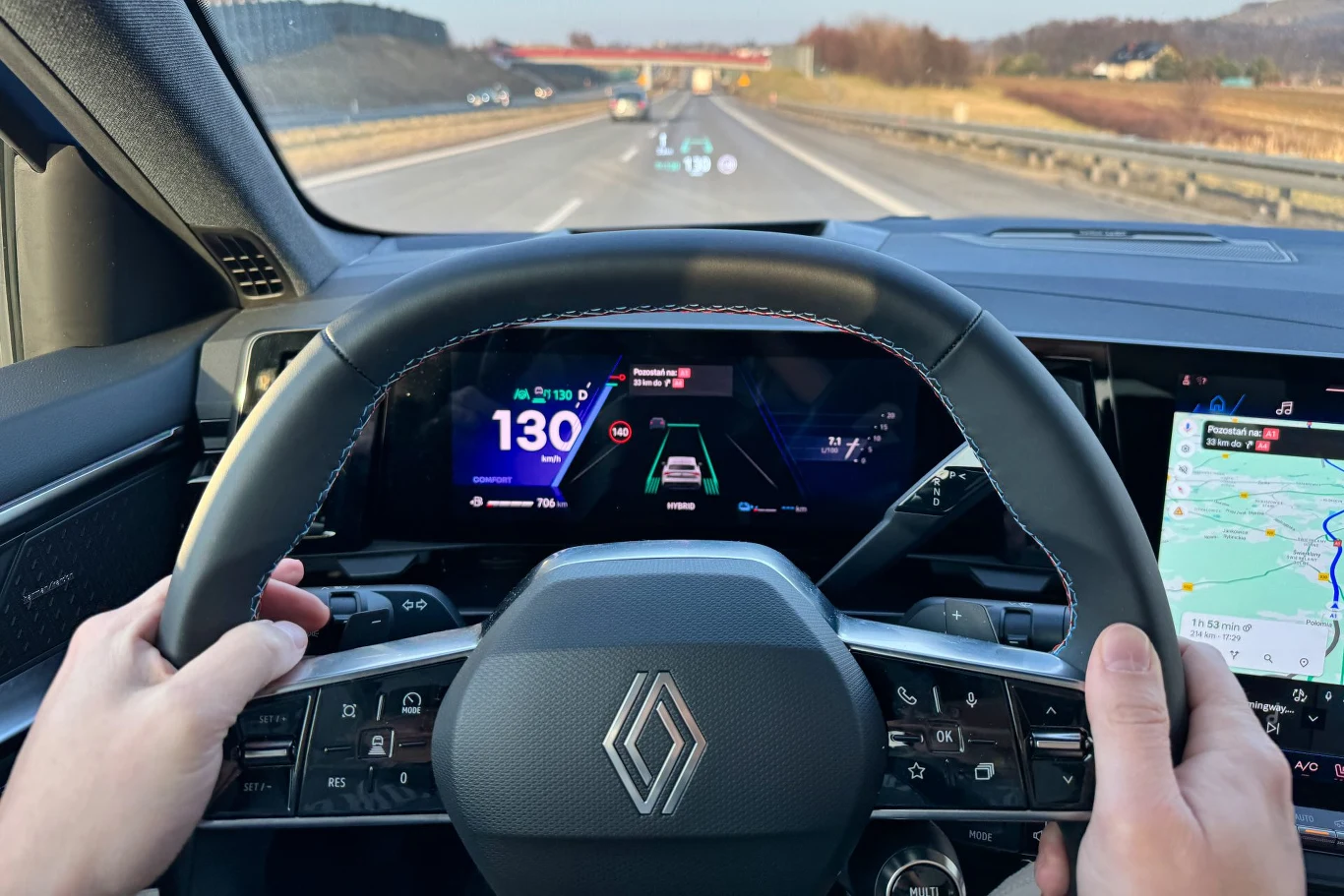 Kierowca na ekranie otrzymuje nawet informacje i położeniu auta na pasie ruchu Widok wnętrza samochodu marki Renault podczas jazdy autostradą, wyświetlacz pokazujący informacje o prędkości oraz trybie jazdy, mapa na ekranie po prawej stronie, dłonie na kierownicy, przednia szyba z wyświetlaczem head-up, widok na drogę i auta prze...