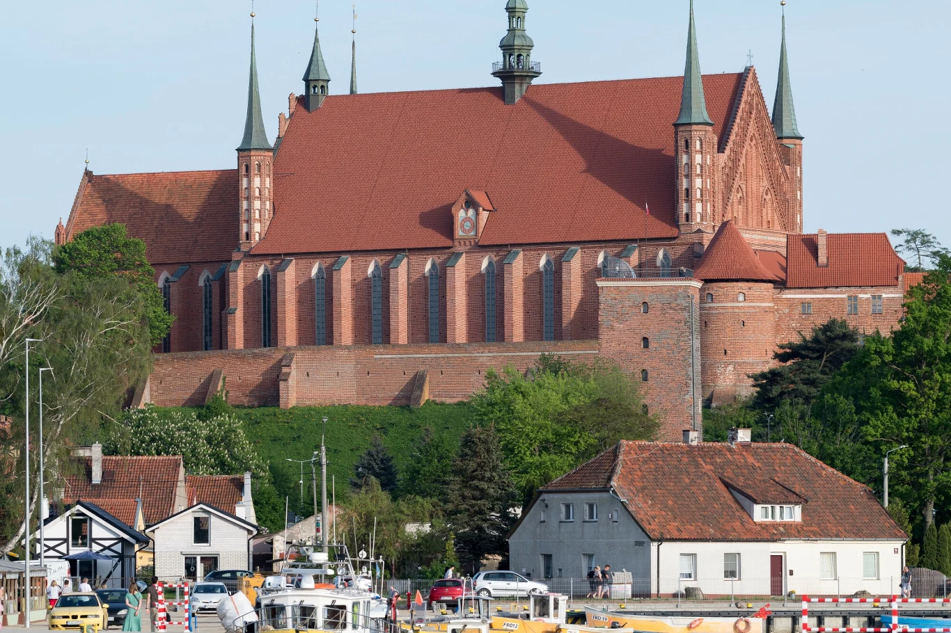 Wzgórze Katedralne i port we Fromborku Ceglany zamek lub katedra z czerwonym dachem i wysokimi wieżami dominuje nad miasteczkiem, w dole widoczne mniejsze domy, przystań z łodziami oraz drzewa w tle.