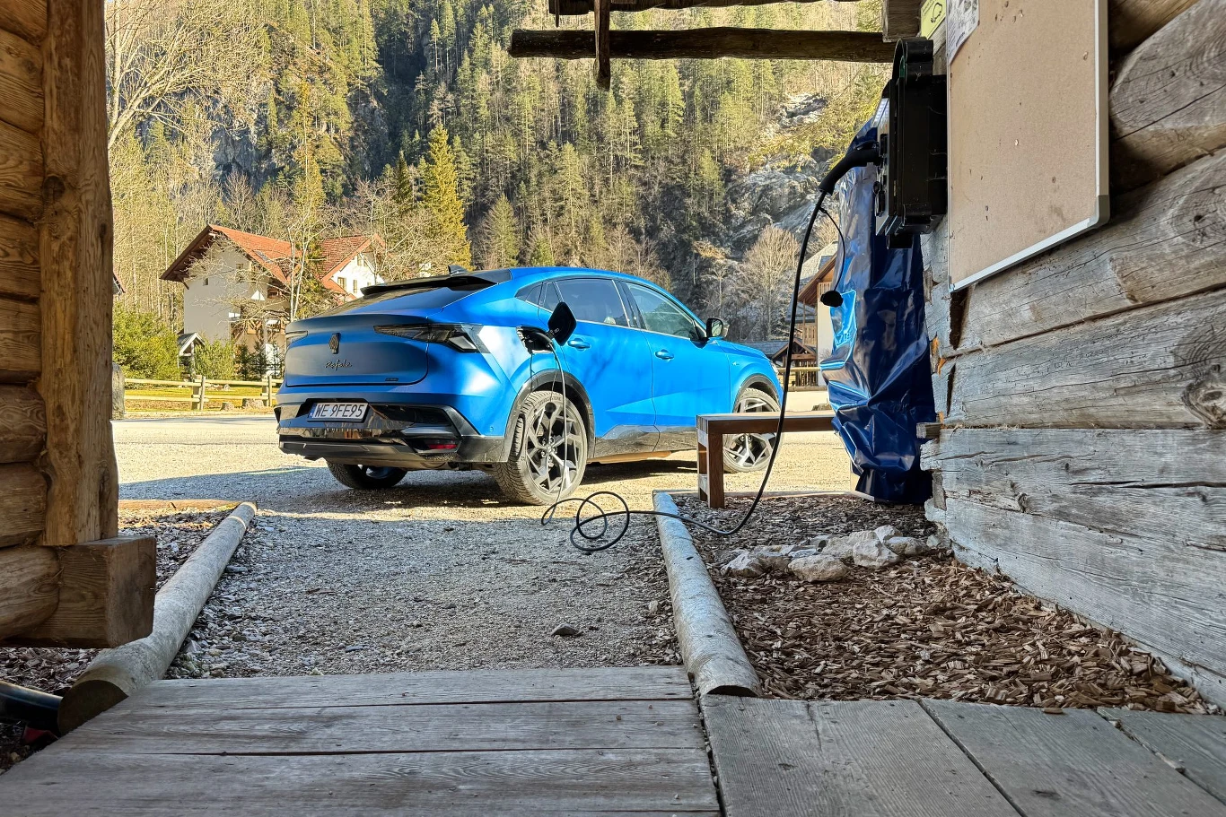 Wersja plug-in może korzystać ze zwykłego gniazdka 230V oraz z ładowarek AC, ładując z mocą do 22 kW Niebieski samochód elektryczny ładowany przy drewnianym budynku, w tle widoczne drzewa oraz górski krajobraz.