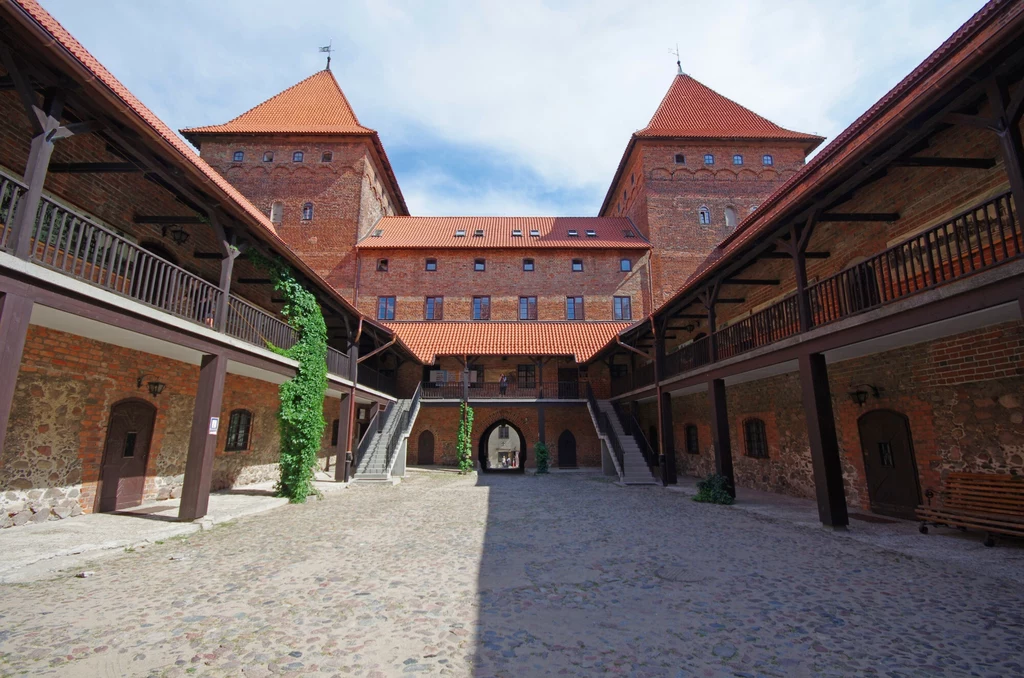 Dziedziniec zamku otoczony ceglanymi murami z arkadami, schodami po obu stronach oraz wejściem z drewnianymi balustradami, centralnie widoczna brama prowadząca do wnętrza budynku.
