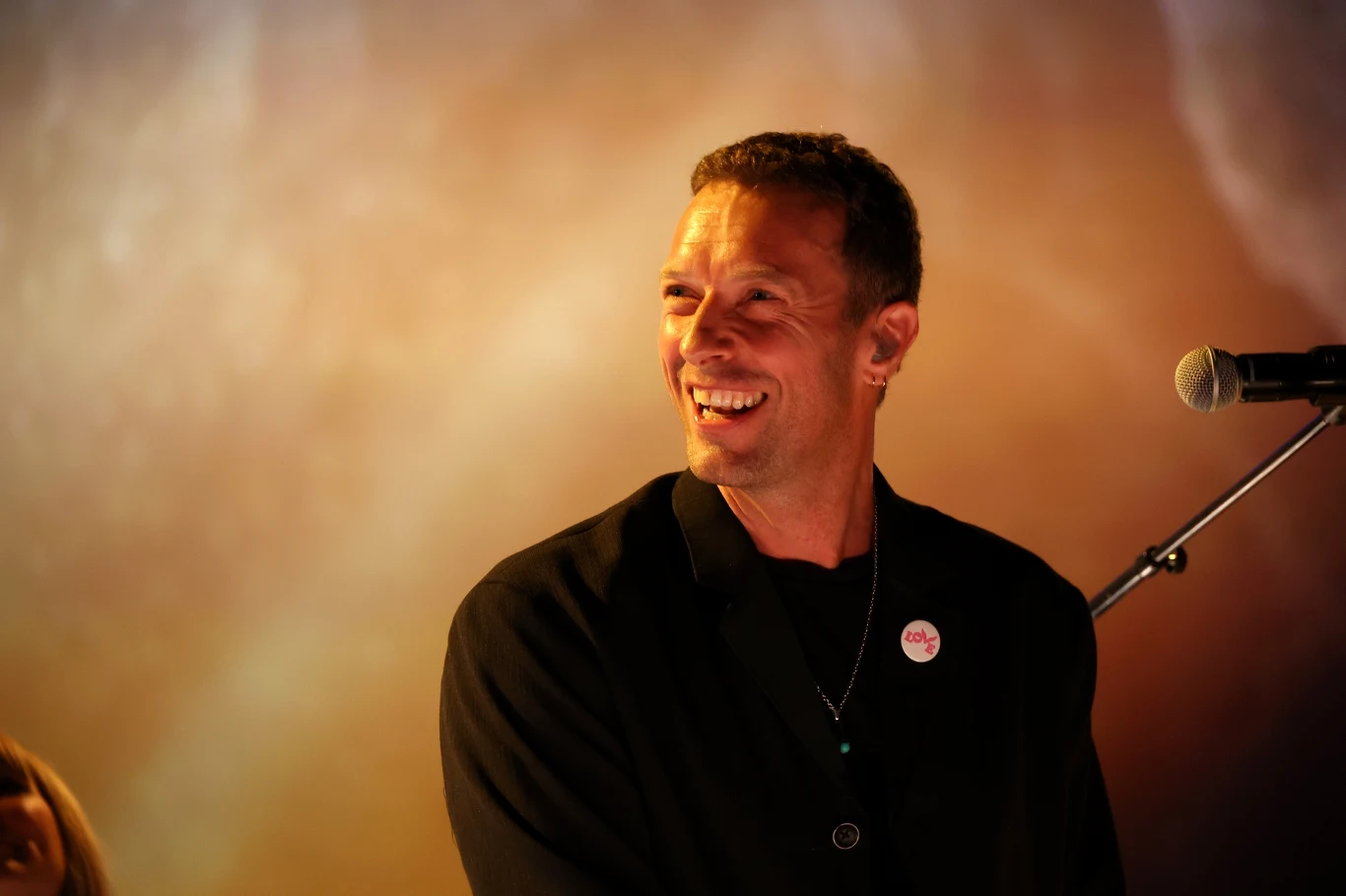 Chris Martin (Coldplay) Uśmiechnięty mężczyzna w czarnej marynarce stoi przy mikrofonie na tle rozmytego, ciepłego światła.