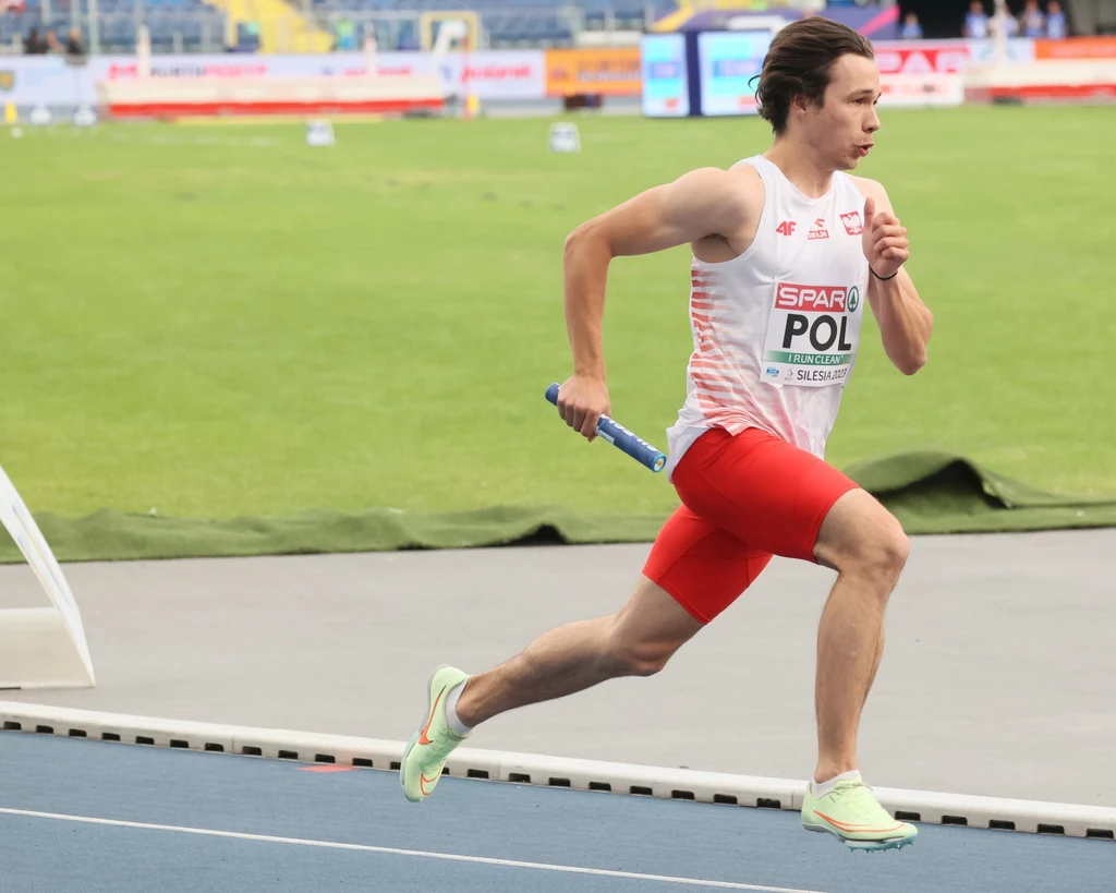 Igor Bogaczyński Lekkoatleta w biało-czerwonym stroju biegnący po bieżni stadionu z pałeczką sztafetową w dłoni, w tle zielona murawa oraz trybuny.