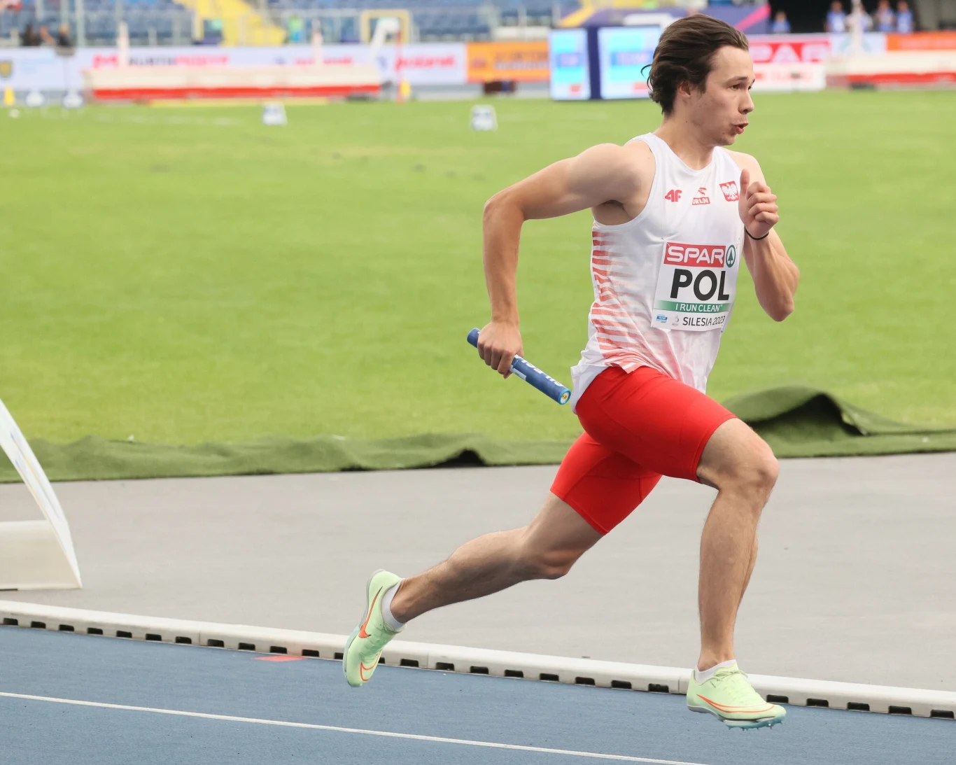 Lekkoatleta w biało-czerwonym stroju biegnący po bieżni stadionu z pałeczką sztafetową w dłoni, w tle zielona murawa oraz trybuny.