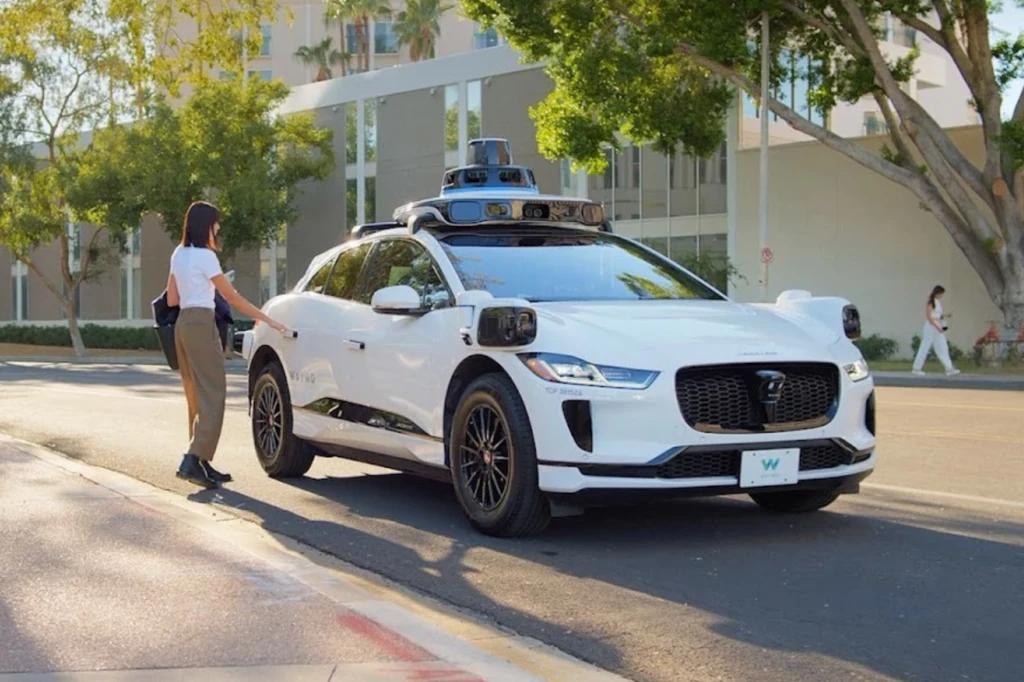 Waymo One to wytwór Alphabet — spółki matki Google. Biała autonomiczna taksówka z zamontowanymi czujnikami i kamerami zatrzymuje się przy krawężniku, a kobieta podchodzi do pojazdu, aby wejść do środka, scena ma miejsce w słonecznym, miejskim otoczeniu z zielenią w tle.