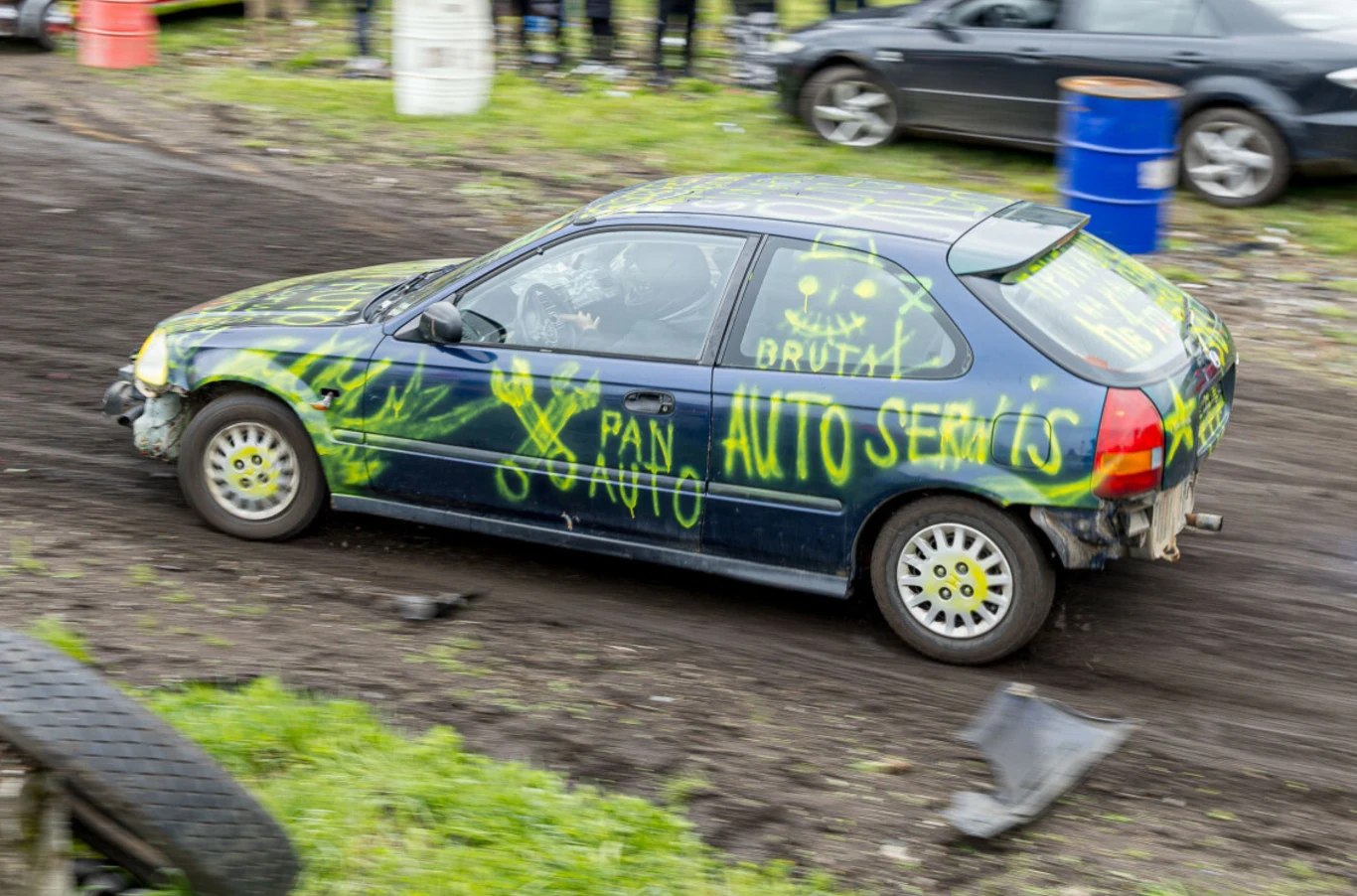 Na Wrak Race w Gliwicach doszło do poważnego zdarzenia (fot. ilustracyjne) Samochód osobowy z wyraźnymi żółtymi napisami oraz rysunkami na karoserii porusza się po nieutwardzonej, błotnistej trasie, wokół widać fragmenty opon, trawę oraz inne pojazdy w tle.