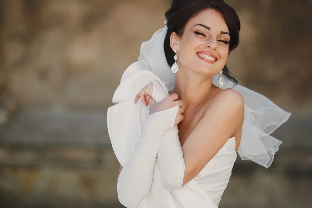 Trend na ślub ze sobą samą przybył już do Polski Uśmiechnięta kobieta w ślubnej sukni i welonie, z eleganckim makijażem oraz ozdobnymi kolczykami, obejmuje się ramionami, prezentując radosny nastrój z jasnym tłem.