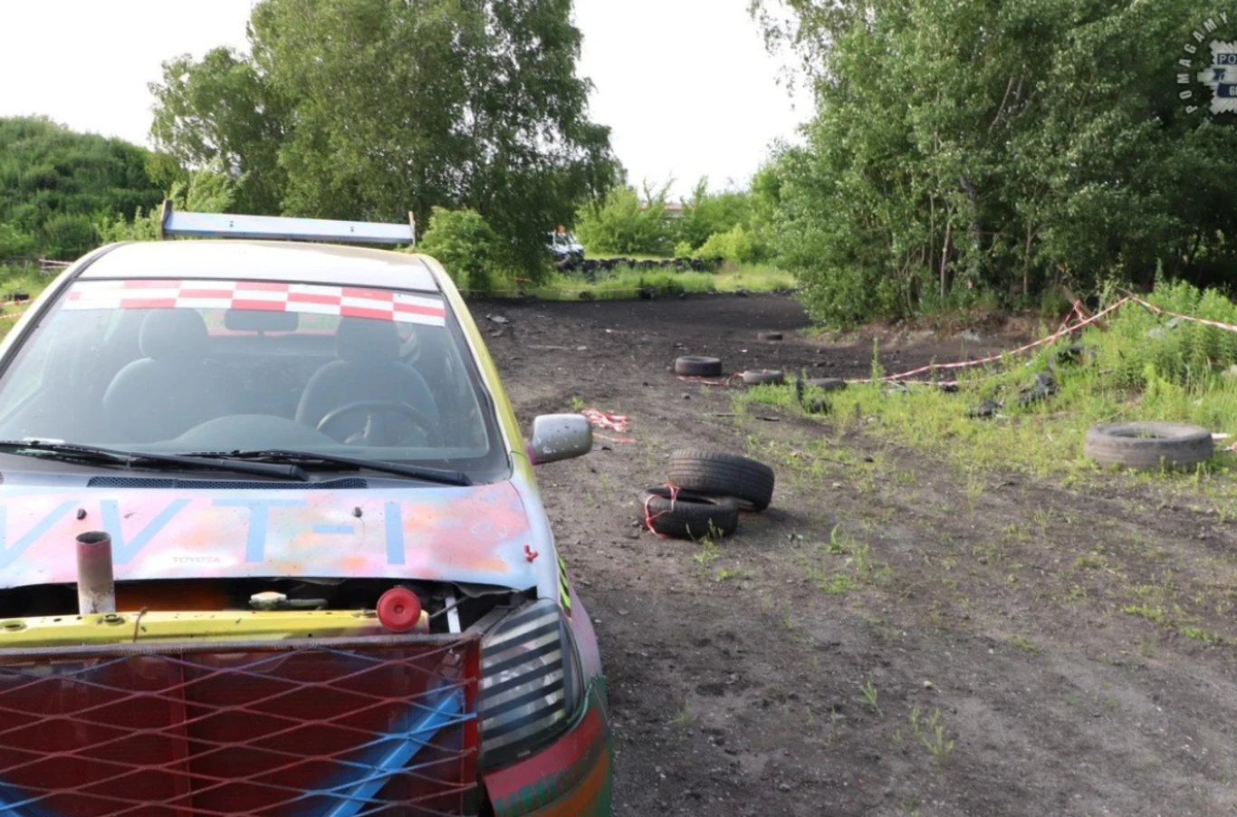 Z Toyoty Yaris odpadło koło i uderzyło stojącą przy trasie kobietę Kolorowy samochód wyścigowy zaparkowany na nieutwardzonym torze z oponami rozrzuconymi wzdłuż trasy oraz taśmą oddzielającą fragment terenu, otoczenie stanowią zieleń i drzewa.