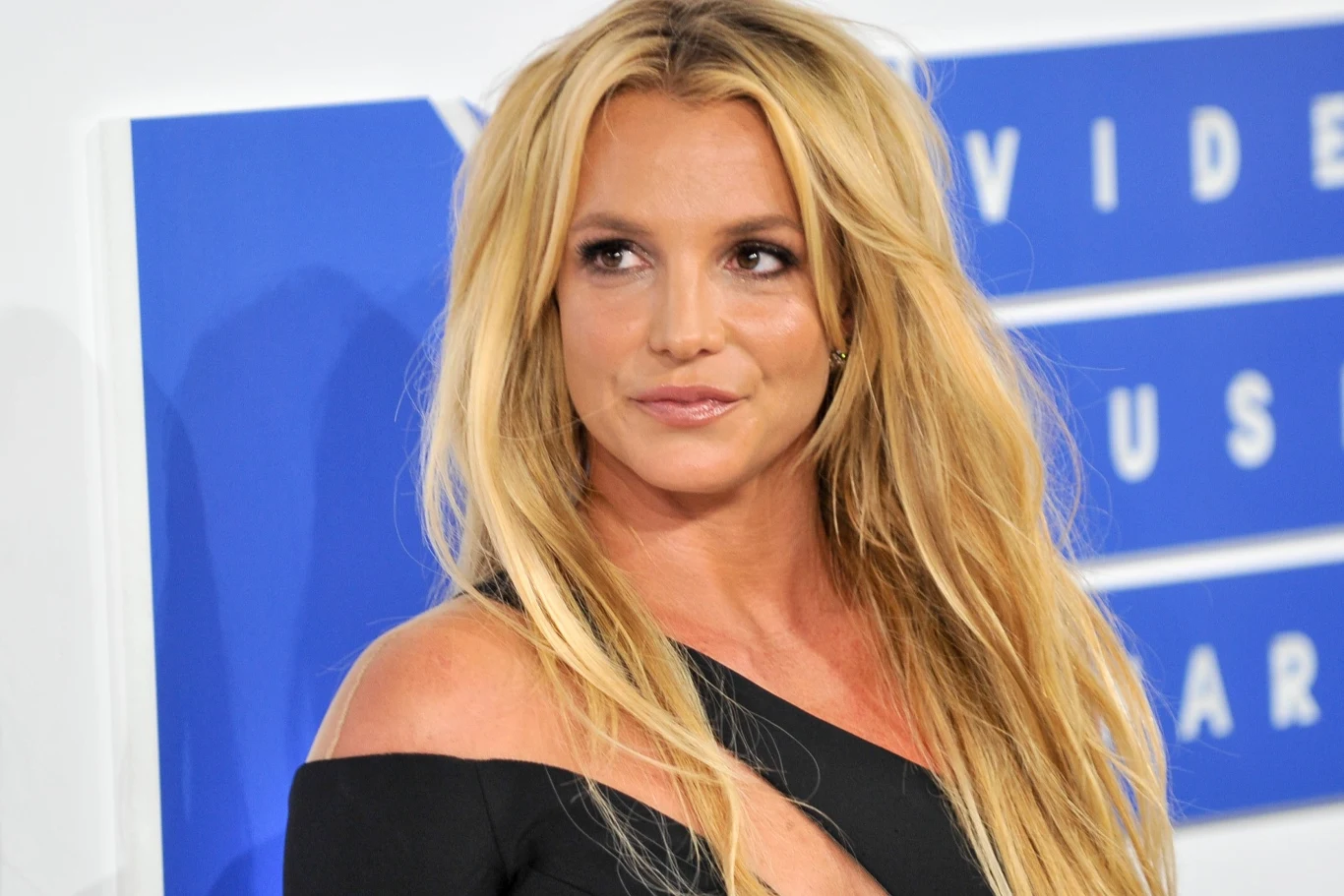 Britney Spears odnawia kontakty z synami Kobieta o długich blond włosach z delikatnym makijażem, ubrana w czarną sukienkę z odkrytym ramieniem, stoi na tle biało-niebieskiej ścianki z napisem związanym z wydarzeniem muzycznym.