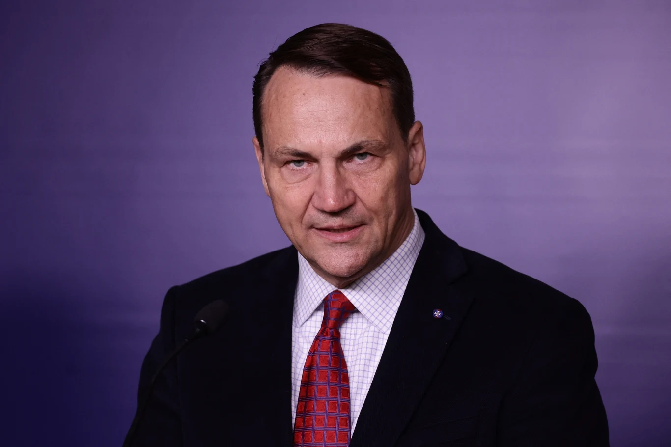 Radosław Sikorski skomentował wygraną Karola Nawrockiego (zdj. ilustracyjne) Mężczyzna w średnim wieku, ubrany w ciemny garnitur, białą koszulę i czerwoną krawat w geometryczny wzór, stojący na tle jednolitej, fioletowej ściany, patrzący prosto w obiektyw.