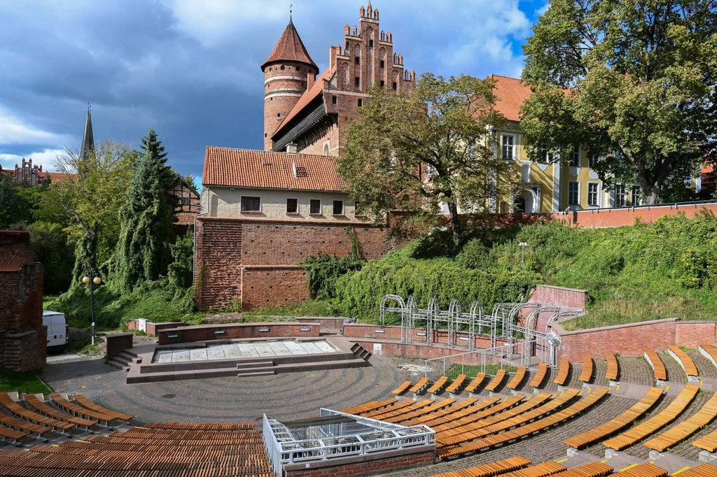 Amfiteatr z półokrągłymi ławkami wykonanymi z drewna i cegły na tle zabytkowego zamku z czerwonej cegły, otoczonego zielenią i drzewami, przy charakterystycznym niebiesko-zachmurzonym niebie.
