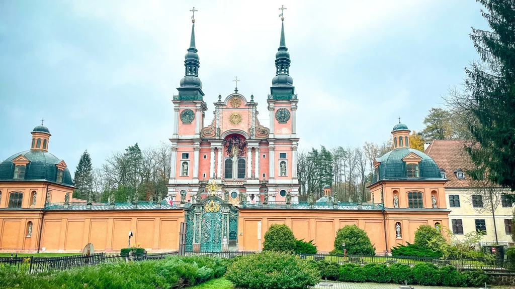Barokowa bazylika z dwoma wysokimi wieżami, otoczona zielenią i ogrodzeniem z misterną bramą, na tle pochmurnego nieba.