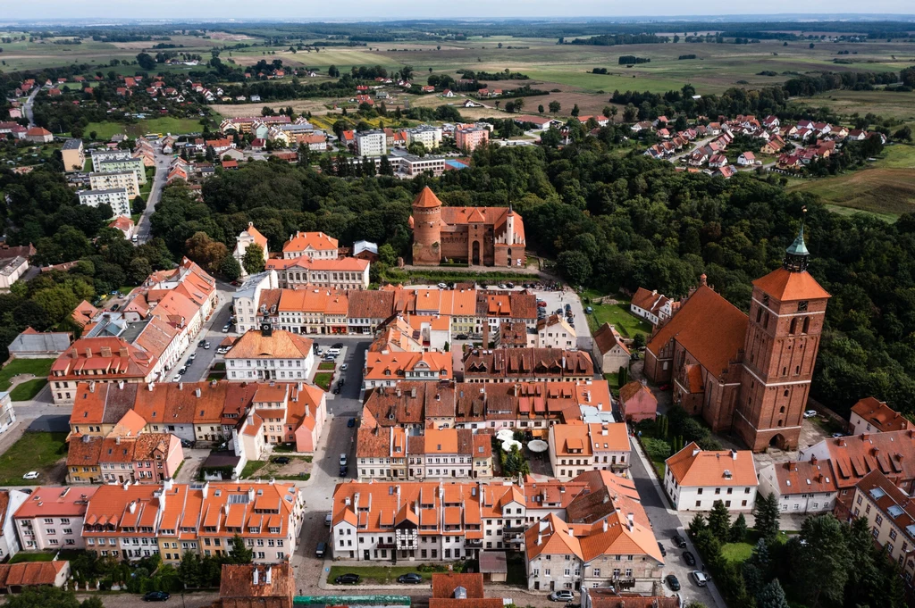 Panorama miasta z czerwonymi dachami, w centrum historyczna zabudowa oraz okazały kościół i zamek otoczone zielenią, w tle osiedla mieszkaniowe oraz rozległe tereny wiejskie.