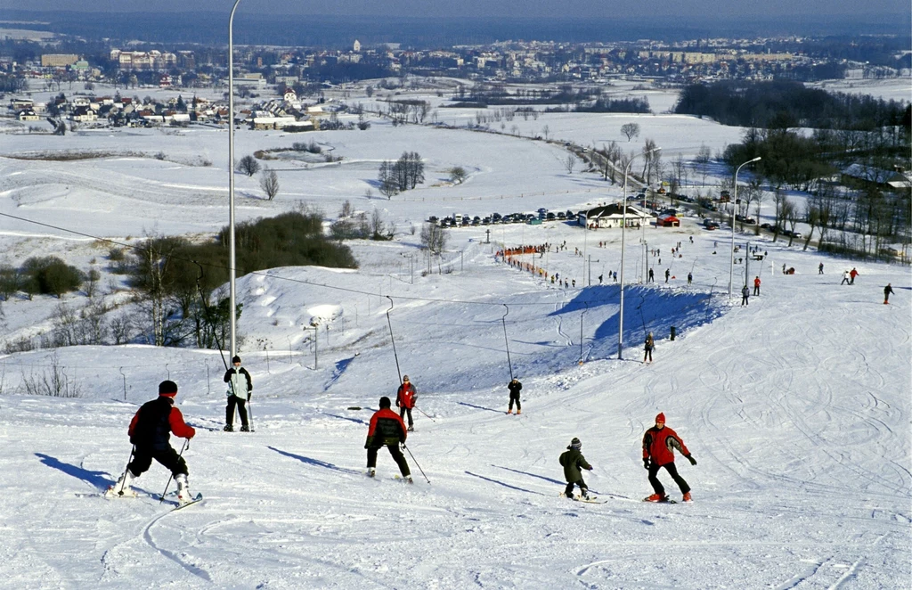 Narciarze zjeżdżający w dół ośnieżonego stoku, oświetlenie narciarskie, w tle rozległa panorama miasta i zimowy krajobraz, na horyzoncie liczne zabudowania i pola pokryte śniegiem.