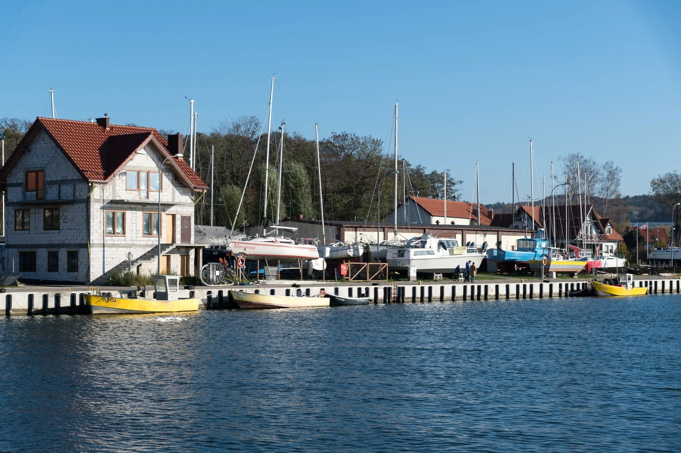 Port rybacki w Tolkmicku nad Zalewem Wiślanym oferuje prawdziwie nadmorski klimat z dala od tłumów, gdzie można obserwować kutry i delektować się świeżą rybą Przystań nad jeziorem z żaglówkami przycumowanymi do pomostu, obok budynki mieszkalne z czerwonymi dachami, na pierwszym planie błękitna woda, całość w słoneczny dzień.