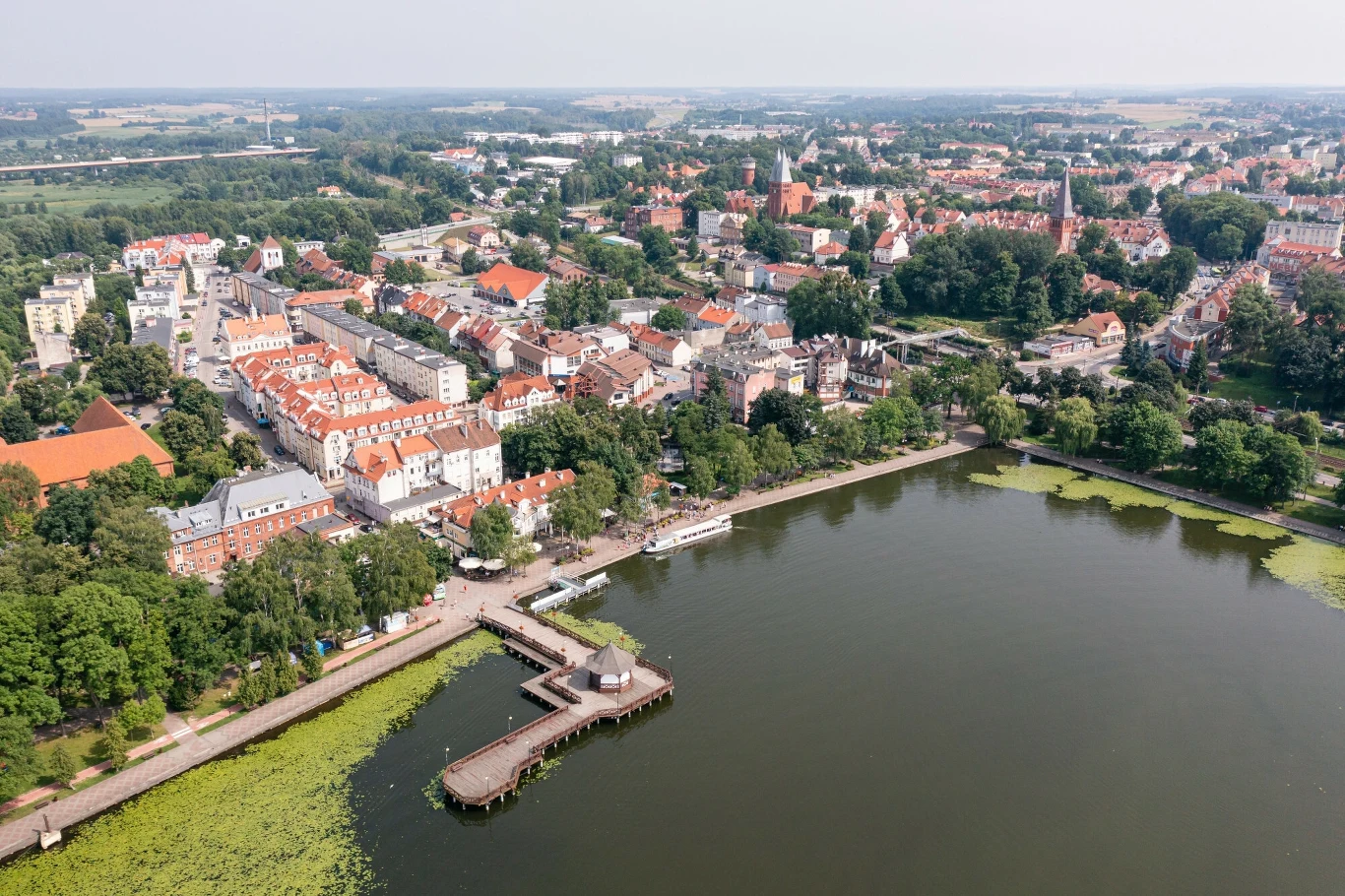 Widok z lotu ptaka na jezioro Drwęckie, molo w Ostródzie oraz miasto z zamkiem krzyżackim Panorama miasta z jeziorem na pierwszym planie, widoczna promenada z drewnianym pomostem wchodzącym w głąb wody, otoczenie stanowią liczne budynki, z przewagą starej zabudowy i zielonych terenów
