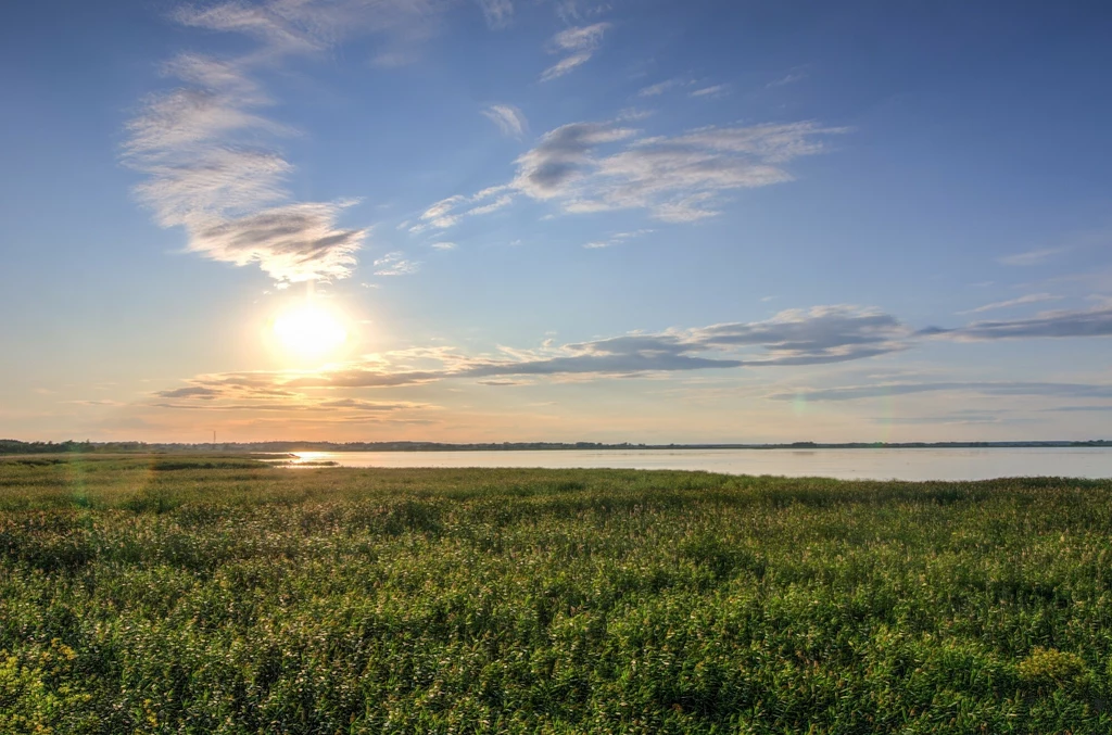 Słońce świeci nad rozległą, zieloną łąką sąsiadującą z jeziorem, a niebo pokrywają rzadkie, lekkie chmury.