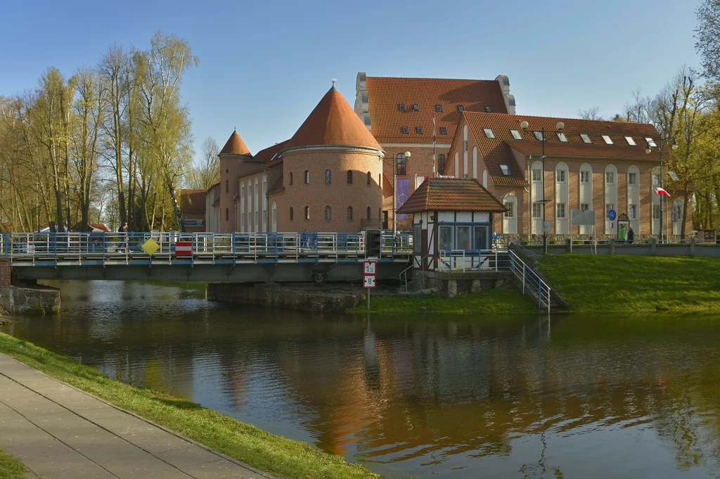Ceglany budynek o okrągłych wieżach i spadzistym dachu położony nad rzeką, widoczny most z barierkami i zielone drzewa w tle, odbicie architektury w spokojnej wodzie.