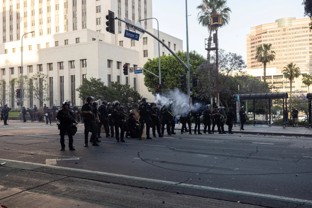 Amerykańscy żołnierze na ulicach Los Angeles. W mieście trwają zamieszki po zatrzymaniu migrantów Grupa policjantów w pełnym wyposażeniu ochronnym rozstawiona na skrzyżowaniu ulic w centrum miasta, jeden z funkcjonariuszy wystrzeliwuje gaz łzawiący, wokół znajdują się wysokie budynki oraz drzewa palmowe, atmosfera napięcia i zamieszania.