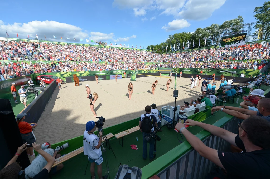 Rozgrywka siatkówki plażowej na stadionie z pełnymi trybunami kibiców, zawodniczki w strojach sportowych, kamera rejestrująca wydarzenie, słoneczna pogoda oraz szerokie, zielone otoczenie stadionu.