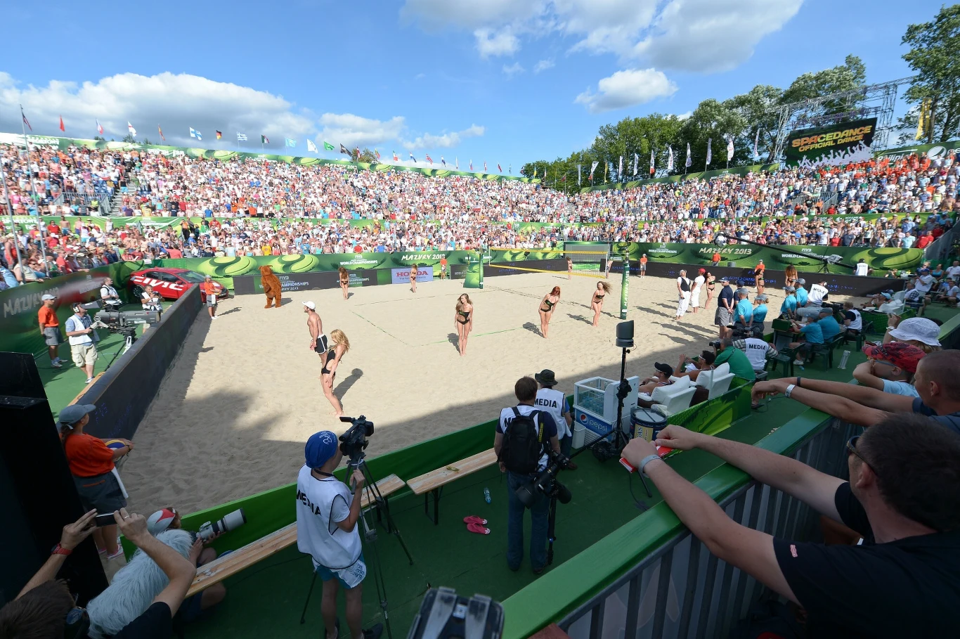 Mistrzostwa Świata w Siatkówce Plażowej w Starych Jabłonkach Rozgrywka siatkówki plażowej na stadionie z pełnymi trybunami kibiców, zawodniczki w strojach sportowych, kamera rejestrująca wydarzenie, słoneczna pogoda oraz szerokie, zielone otoczenie stadionu.