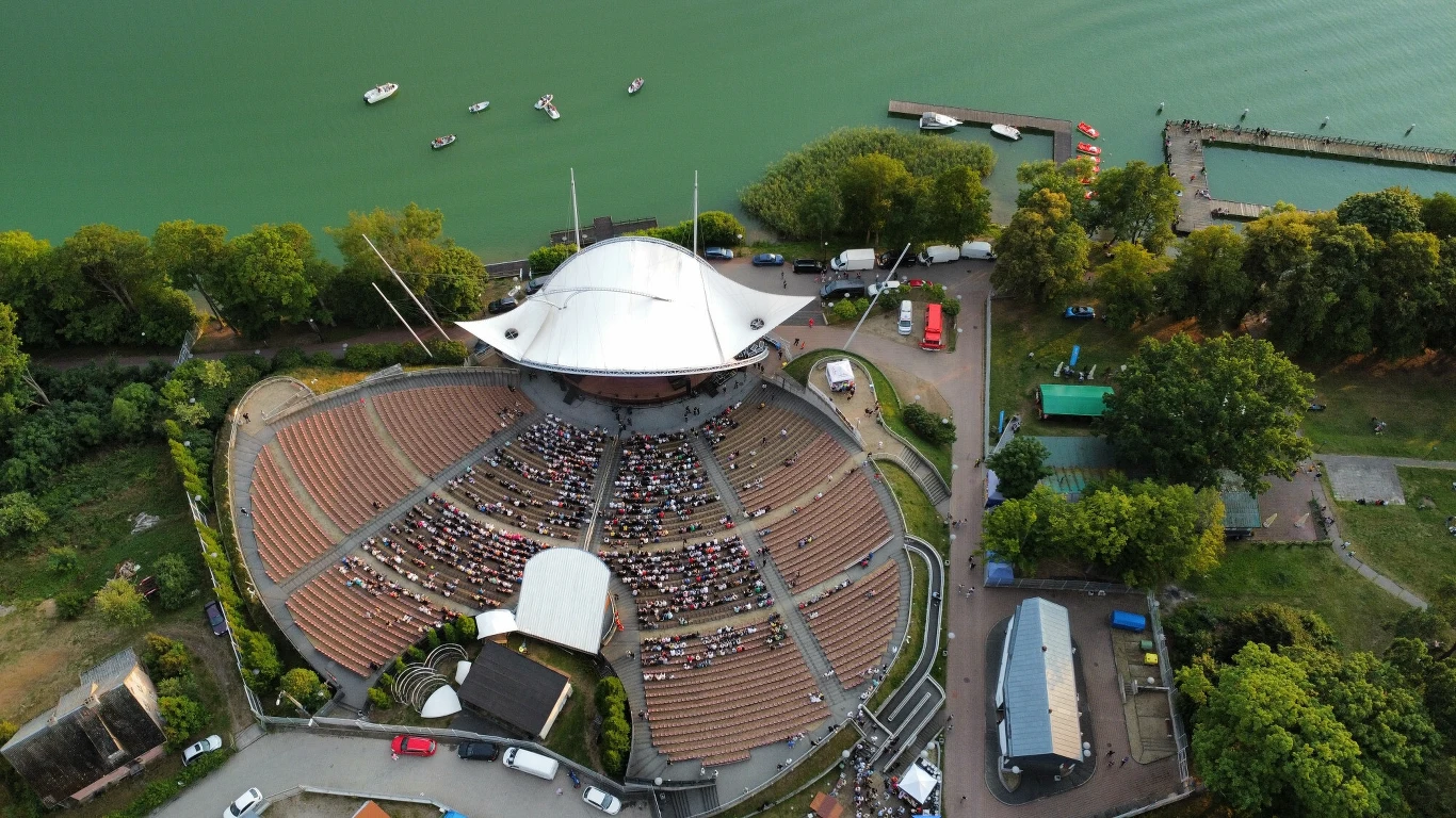 Amfiteatr nad jeziorem Czos w Mrągowie, gdzie odbywają się liczne festiwale Amfiteatr z zadaszoną sceną i schodkowymi trybunami, wypełnionymi publicznością, położony nad jeziorem, otoczony zielenią oraz kilkoma budynkami i drogą, na wodzie widoczne łódki.