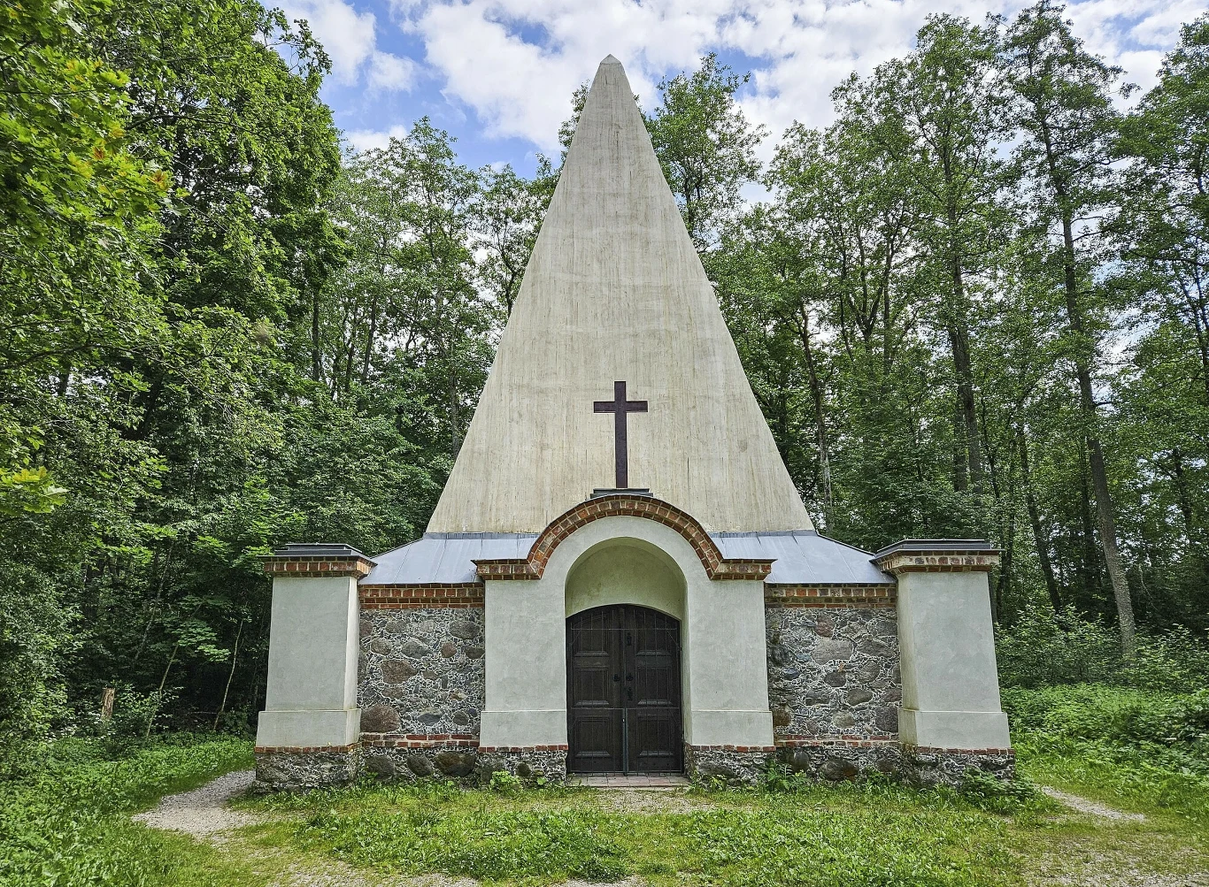 Piramida w Rapie. Kaplica i grobowiec rodzinny pruskiego rodu baronów von Fahrenheid Kaplica o unikalnej, trójkątnej konstrukcji dachu z dużym krzyżem na froncie, zlokalizowana w otoczeniu zielonych, wysokich drzew, z murowaną kamienno-ceglaną podstawą oraz dużymi, ciemnymi drzwiami wejściowymi.