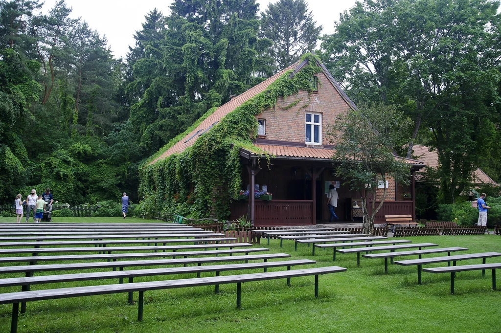 Drewniane ławki ustawione rzędami na zielonej trawie przed budynkiem z czerwonej cegły porośniętym bluszczem, otoczonym wysokimi drzewami, kilka osób spaceruje wokół domu.