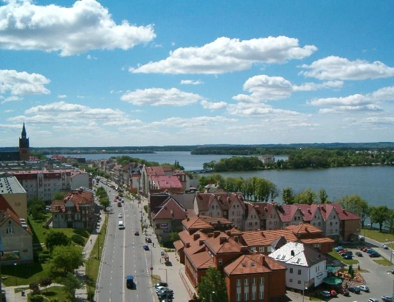 Ełk: dynamiczne miasto nad jeziorem, gdzie miejski zgiełk spotyka się z mazurskim spokojem Panorama miasta z widokiem na jezioro, liczne czerwone dachy domów, szeroka ulica prowadząca w stronę wody, w tle widoczny kościół z wysoką wieżą i rozciągające się zielone tereny.