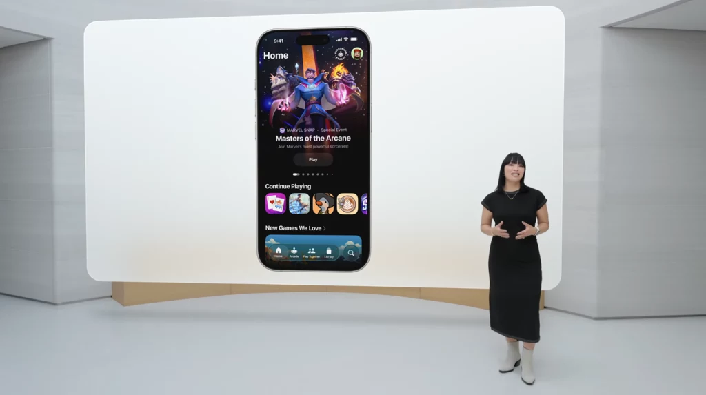 Gry to nowa aplikacja z iOS 26. Duży ekran prezentuje interfejs aplikacji na smartfonie z grą Masters of the Arcane, przed ekranem stoi kobieta w czarnej sukience wygłaszająca prezentację.