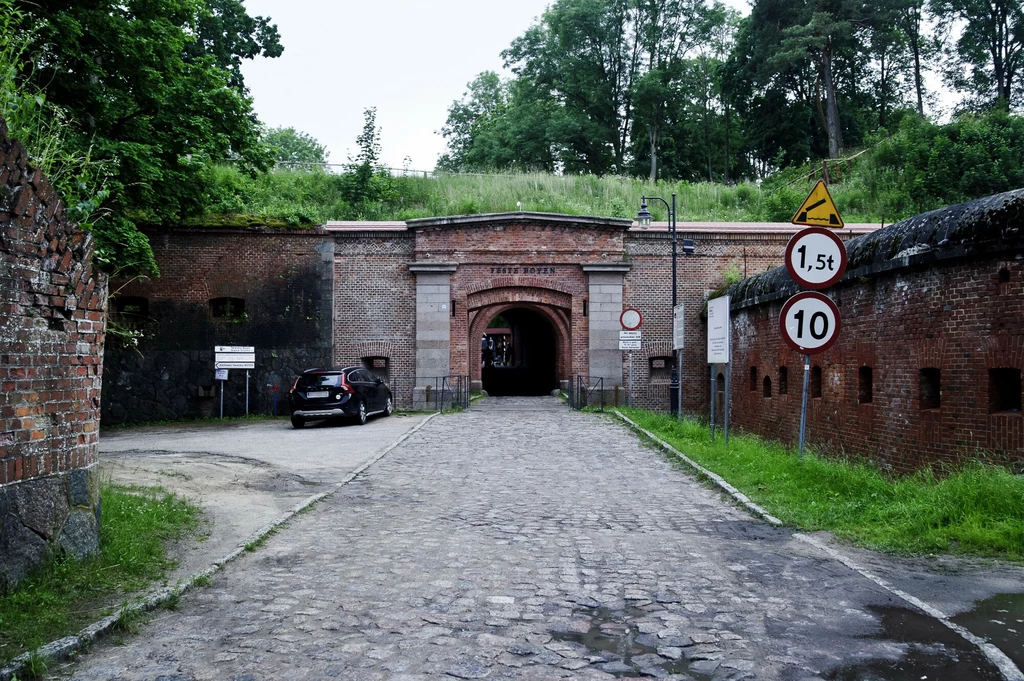 Ceglany fort z łukowym przejazdem, przed którym znajduje się brukowana droga, znak ograniczenia prędkości oraz zakaz wjazdu pojazdów powyżej 1,5 tony, po lewej stronie zaparkowany samochód, otoczenie z bujną zielenią i drzewami.