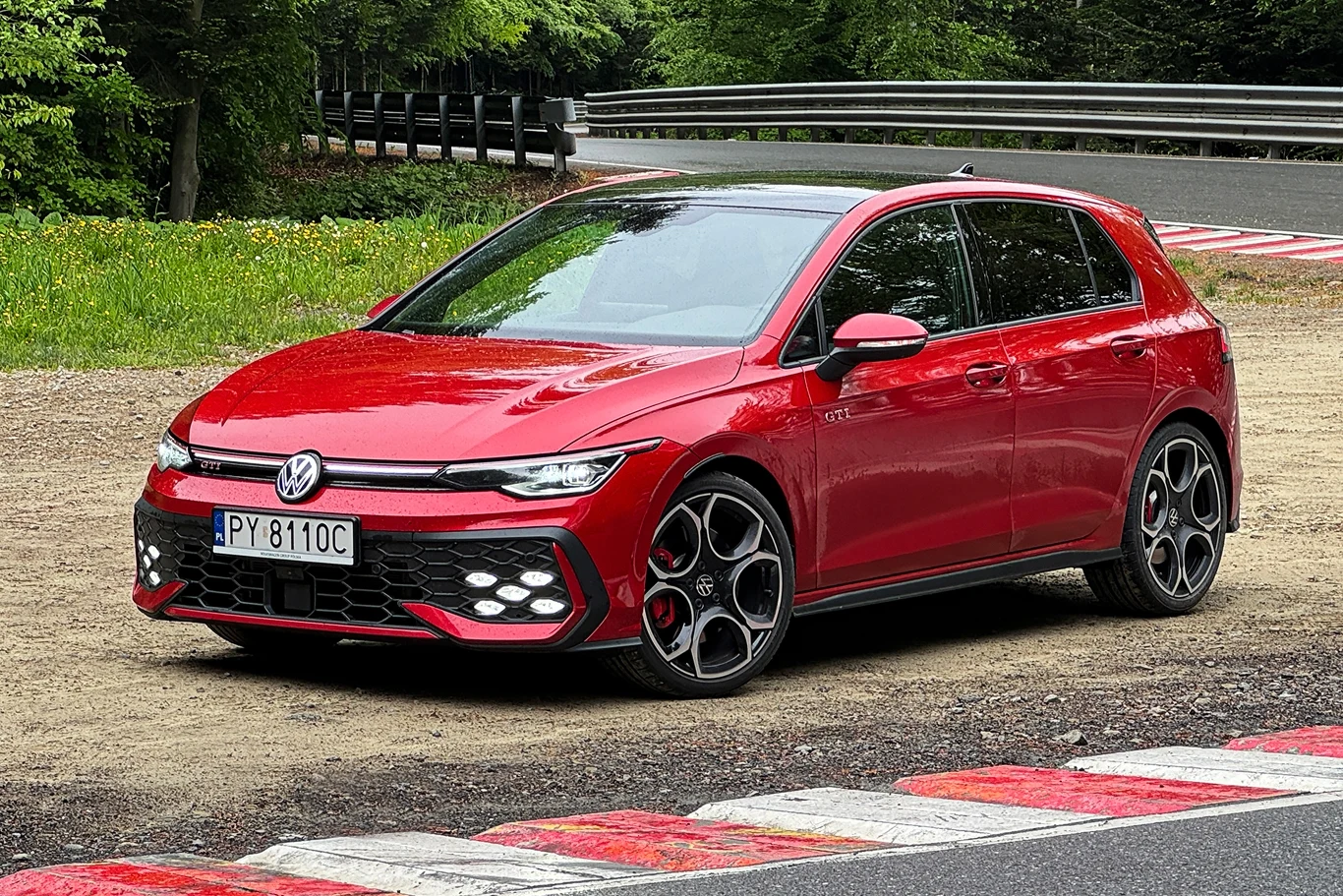 Volkswagen Golf GTI po liftingu. Mocniej, szybciej i bez wad poprzednika Czerwony samochód marki Volkswagen Golf GTI stoi przy torze wyścigowym, na tle zielonych drzew i zakrętu z barierami ochronnymi, światła samochodu odbijają się w lakierze, widoczna rejestracja.
