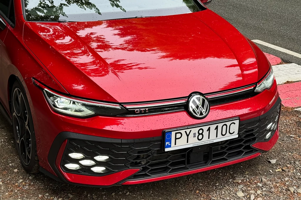 Modyfikacje zewnętrzne objęły między innymi lekko przeprojektowany przedni zderzak. Czerwona sportowa osobówka marki Volkswagen GTI z widoczną przednią atrapą chłodnicy, reflektorami LED oraz polską tablicą rejestracyjną, pojazd stoi na poboczu obok asfaltowej drogi.