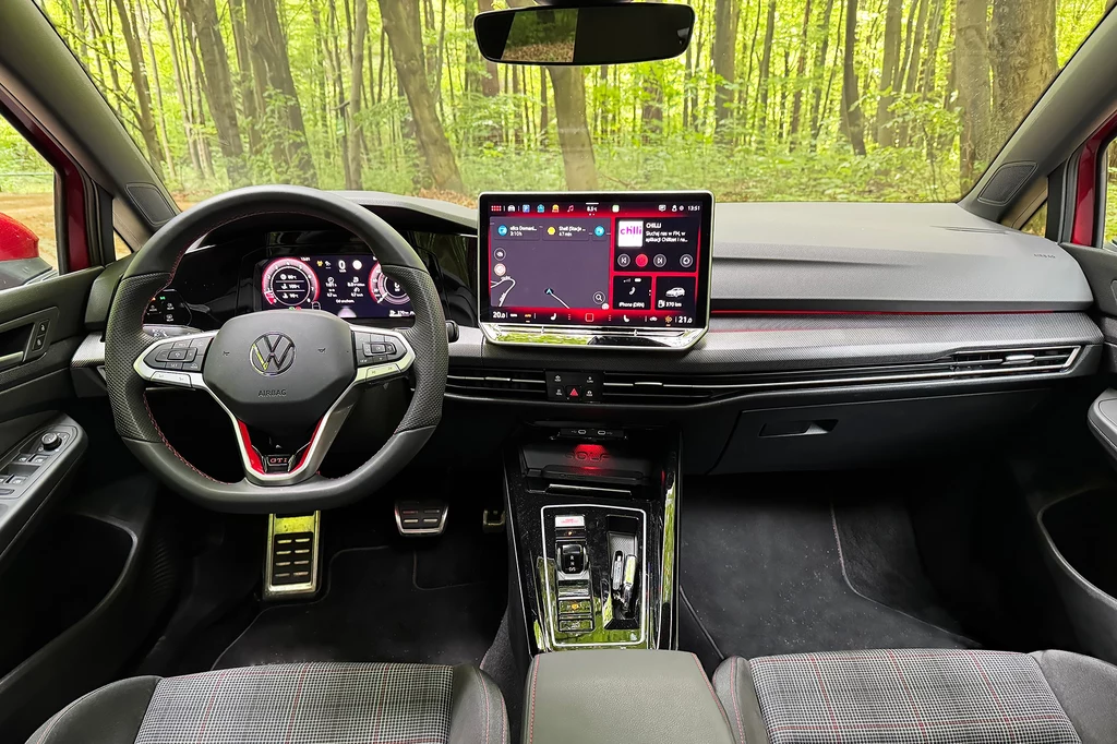 Więcej zmian zauważymy we wnętrzu. Nowością jest m.in. system multimedialny w formie tabletu. Nowoczesne wnętrze samochodu marki Volkswagen z cyfrowym zestawem wskaźników, dużym ekranem dotykowym pośrodku deski rozdzielczej oraz sportową kierownicą. Tło stanowi widok lasu za szybą.