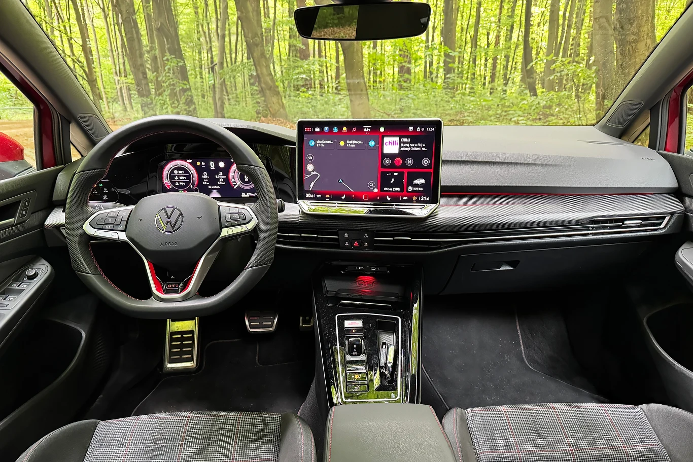 Więcej zmian zauważymy we wnętrzu. Nowością jest m.in. system multimedialny w formie tabletu. Nowoczesne wnętrze samochodu marki Volkswagen z cyfrowym zestawem wskaźników, dużym ekranem dotykowym pośrodku deski rozdzielczej oraz sportową kierownicą. Tło stanowi widok lasu za szybą.
