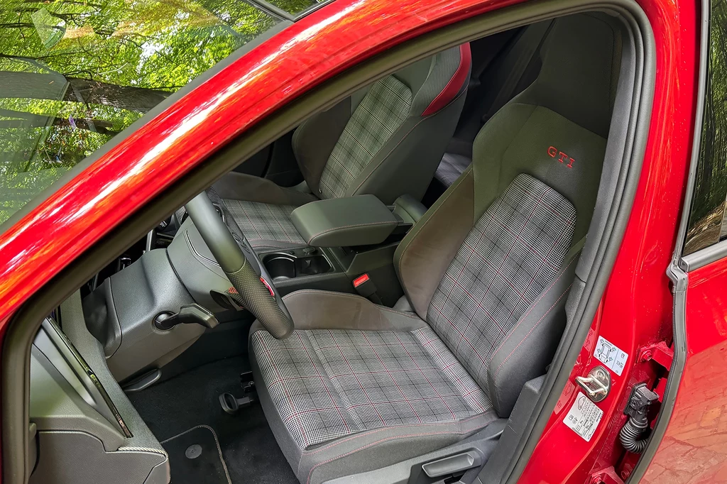 Fotele w kratę to znak szczególny Golfów GTI. Taką tapicerkę miały już pierwsze modele. Wnętrze czerwonego samochodu z dobrze widocznym przednim fotelem obszytym materiałem w kratkę oraz napisem GTI na oparciu, z elementami konsoli środkowej i kierownicą po lewej stronie.