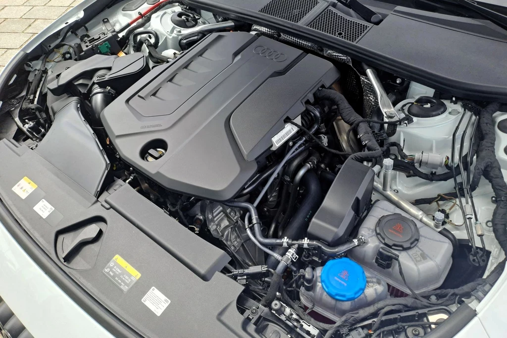 Audi A5 wyposażone jest w dwulitrowy silnik benzynowy o mocy 251 KM. Współpracuje on z układem elektrycznym generującym 142 KM. Komora silnika samochodu marki Audi z widoczną jednostką napędową, przewodami oraz elementami układu chłodzenia i płynów eksploatacyjnych.