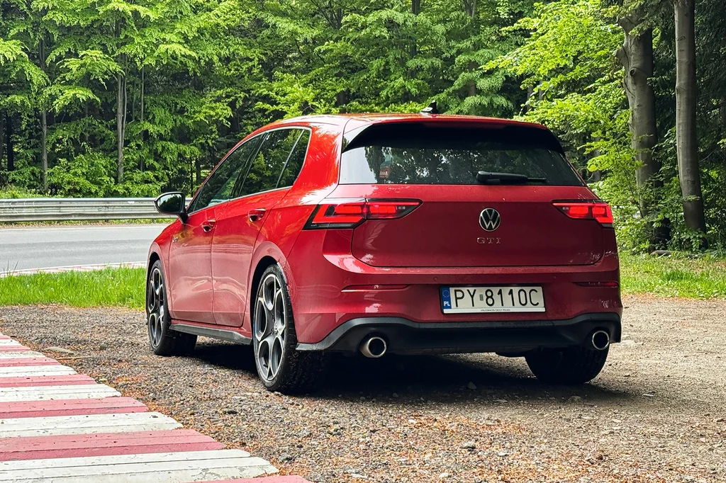 Wrażenia z jazdy potęguje adaptacyjne zawieszenie DCC, które oferuje aż 15 różnych ustawień. Czerwony samochód marki Volkswagen Golf stoi zaparkowany na poboczu przy drodze otoczonej lasem, widoczne sportowe felgi i detale modelu GTI, auto skierowane tyłem.